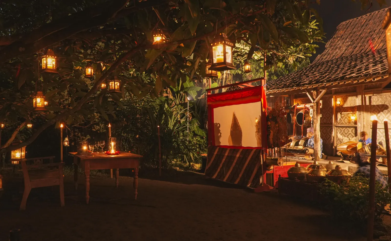 A candlelit table under lanterns beside a shadow-puppet screen at night