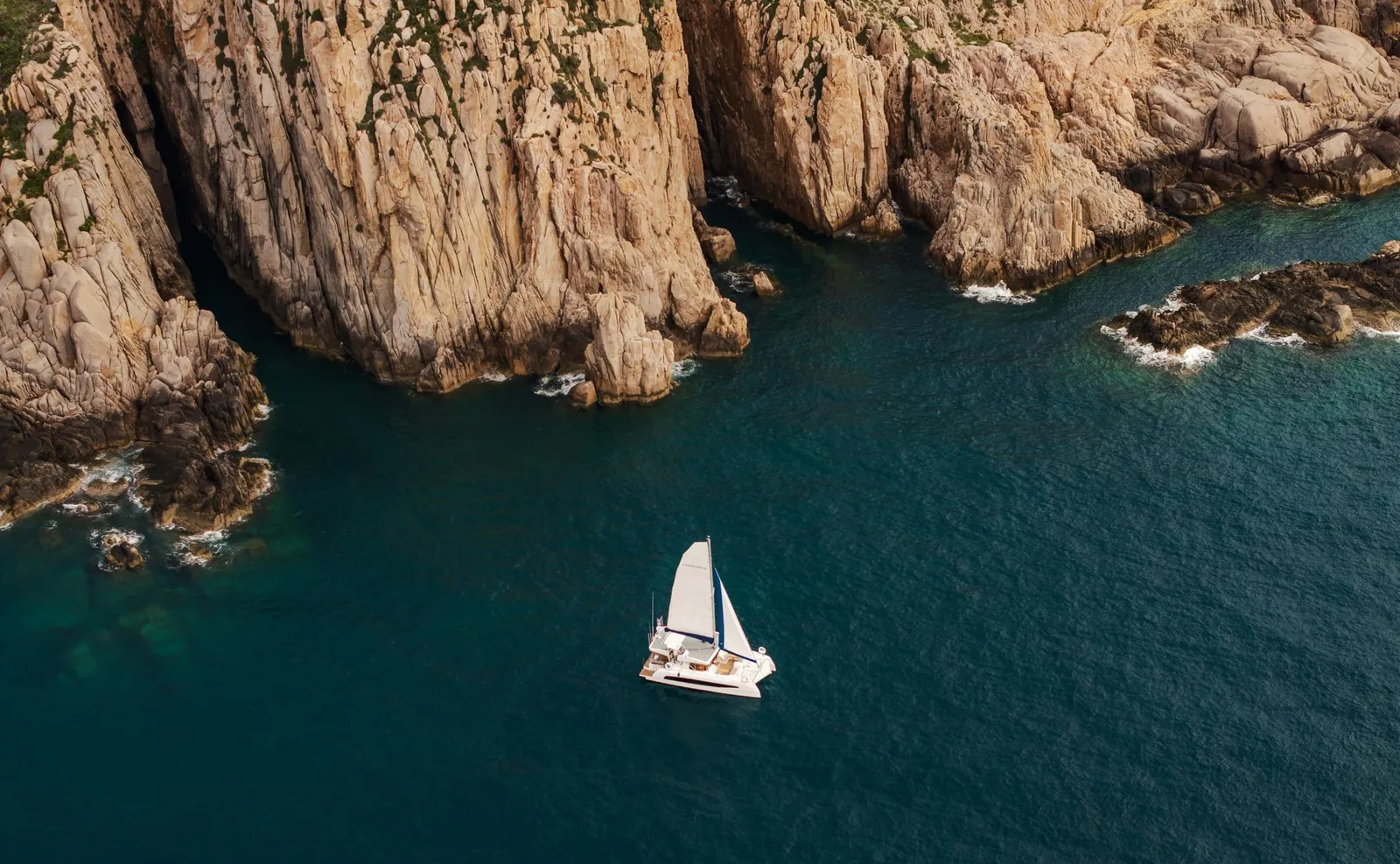 The white catamaran anchored in a cove between the rocks of the coast, seen from above