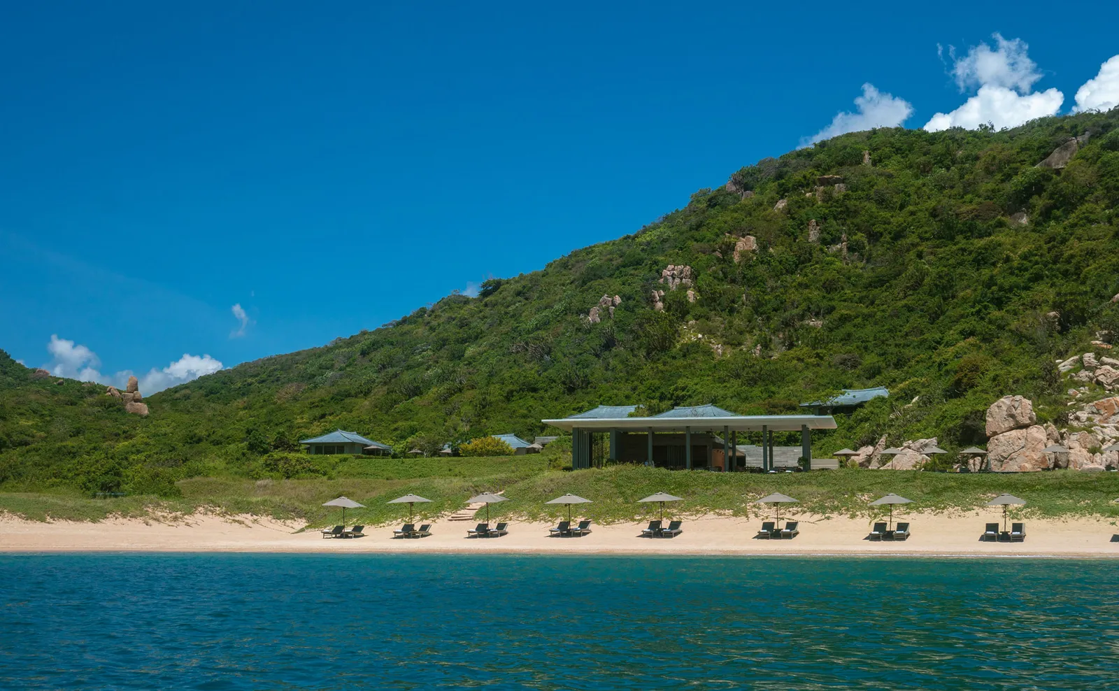 The private beach from the water: loungers on the pale sand, the Beach Club and the wooded hill behind