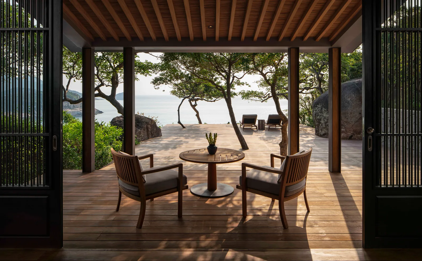 An Ocean Pavilion deck: 2 armchairs and a table between the sliding doors, the sea beyond the trees