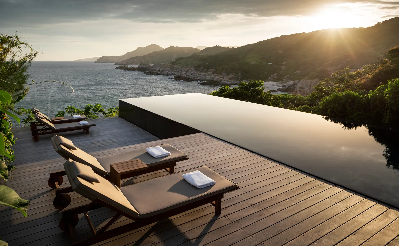 Loungers on the timber deck beside the infinity pool, with the coast at sunset