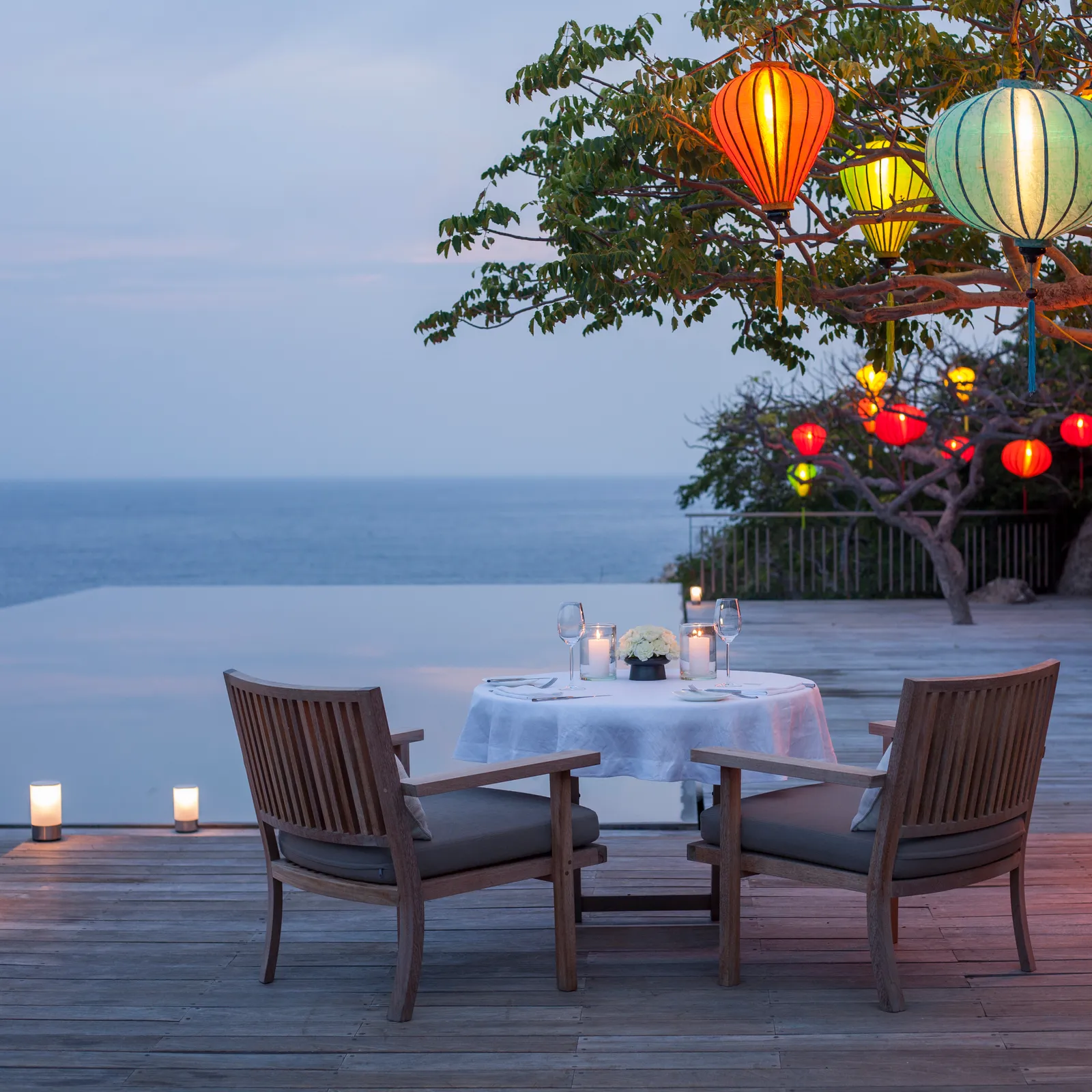 A table laid on the Cliff Pool deck at dusk, lanterns hanging from the tree and the sea below