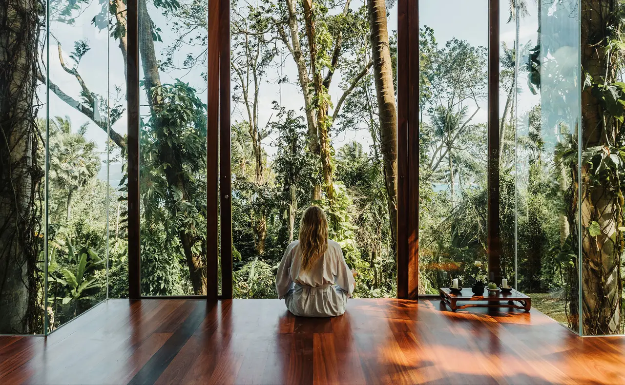 A guest sitting in a glass-walled pavilion on a wooden floor, the forest outside