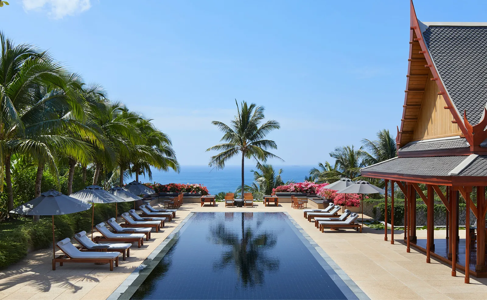 A long villa pool aimed at the sea between red-roofed pavilions