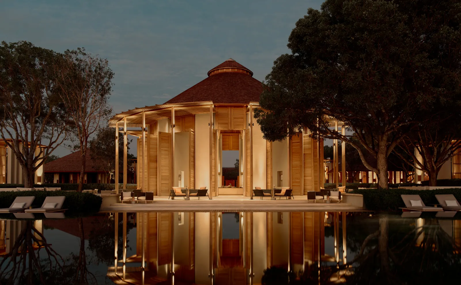 The bar pavilion lit at dusk, reflected in the pond, with loungers on the terrace and the big trees.