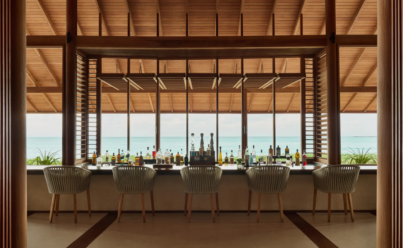 The Beach Club’s bar under its timber roof: a line of stools, a row of bottles and the sea beyond the glass.