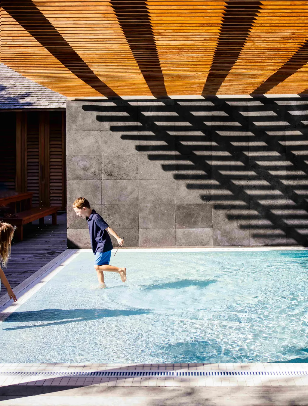 A child runs through the shallow water of a pool, under a slatted timber canopy casting stripes of shade.