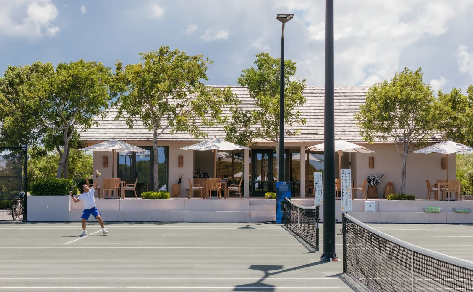A player serves on a clay court before the Clubhouse, parasols and trees behind the net.
