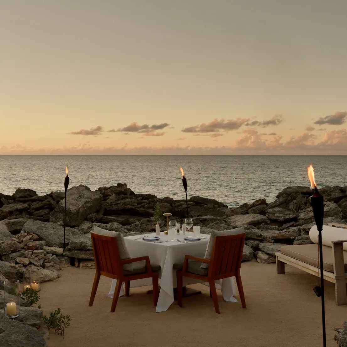 A table for 2 laid on the shore rock at sunset, between lit torches, facing the sea.