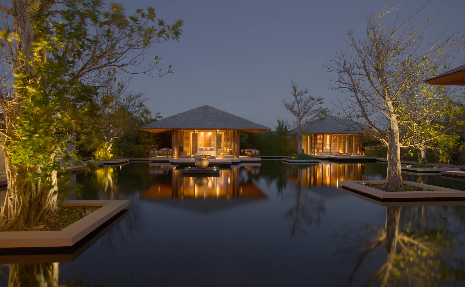 2 pavilions lit at night, reflected in a villa’s still pond, trees in silhouette.