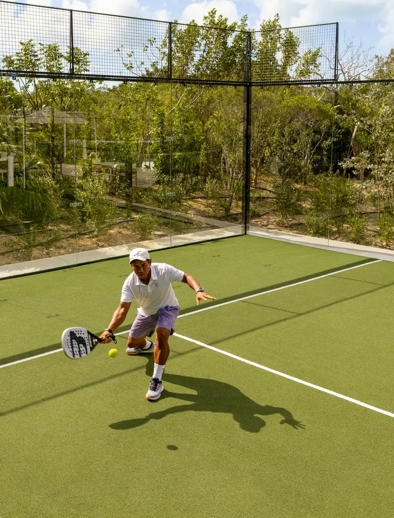 A padel player reaches for a low ball on a green court, the fence and the scrub behind.