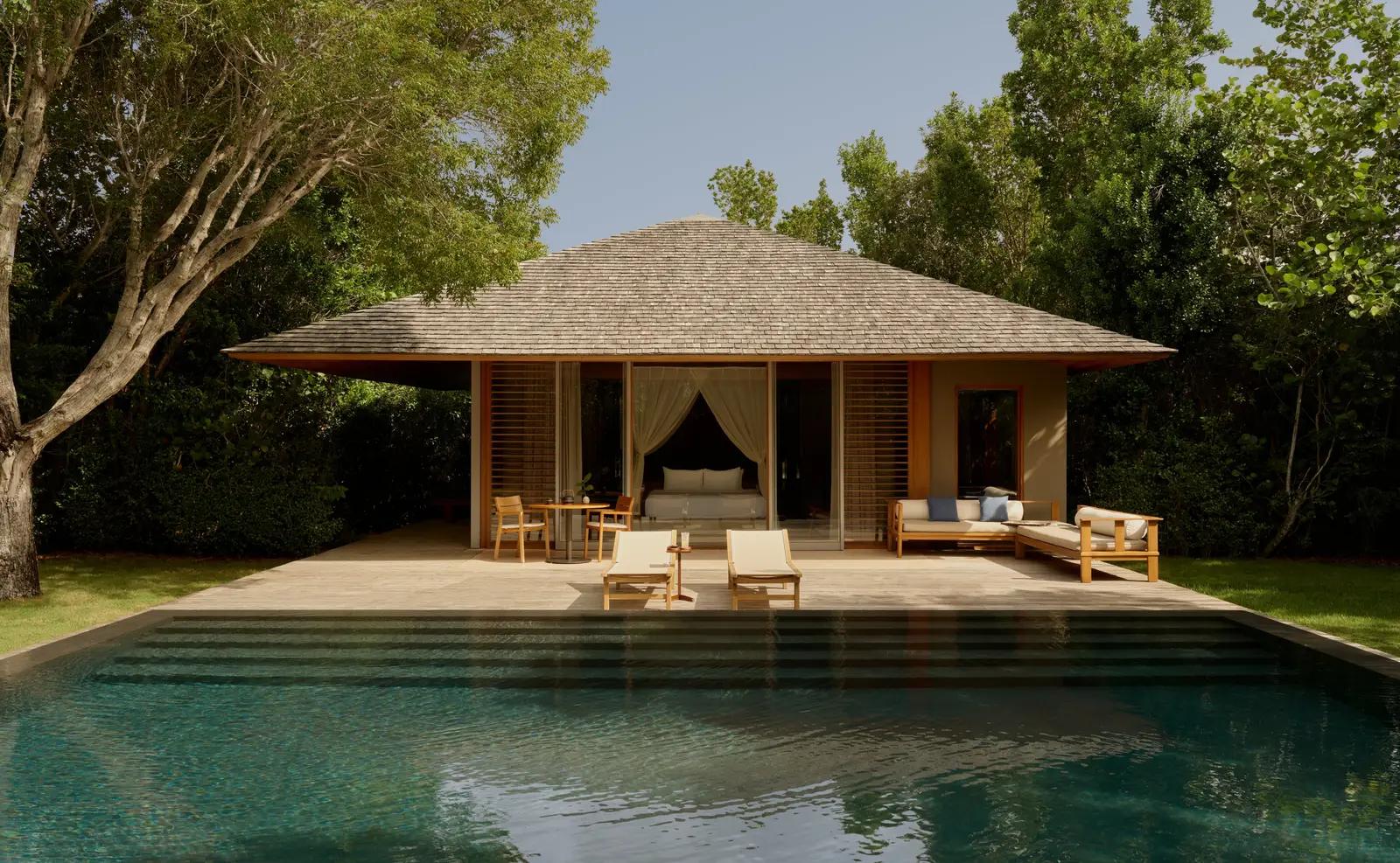 An Alcove Pool Pavilion beyond its green pool: the bed behind glass, loungers and a sofa on the deck.