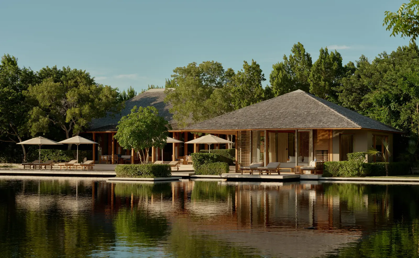 A 4-bedroom villa seen across its pond: pavilions, parasols and trees reflected in still water.