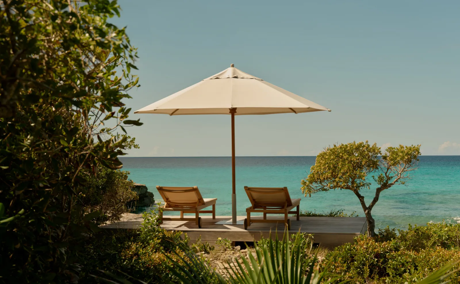 2 timber loungers under a pale parasol, on a platform among shrubs above the turquoise sea.