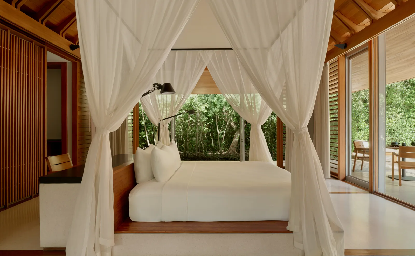 An Ocean Pavilion’s bed under its white canopy, with a reading lamp and vegetation beyond the glass.