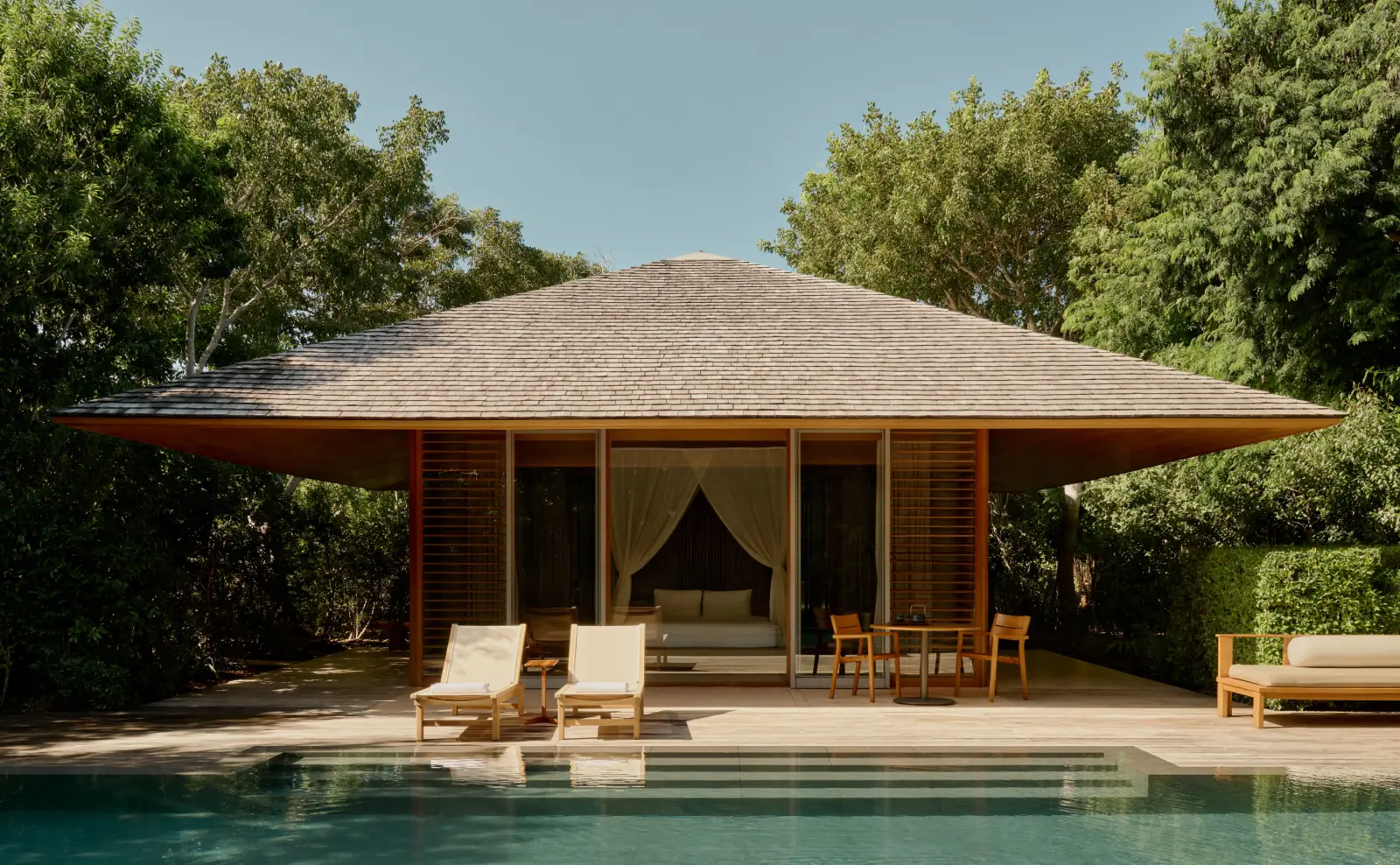 A Pool Pavilion seen from its pool: the wide hipped roof, 2 loungers and a table on the timber deck.