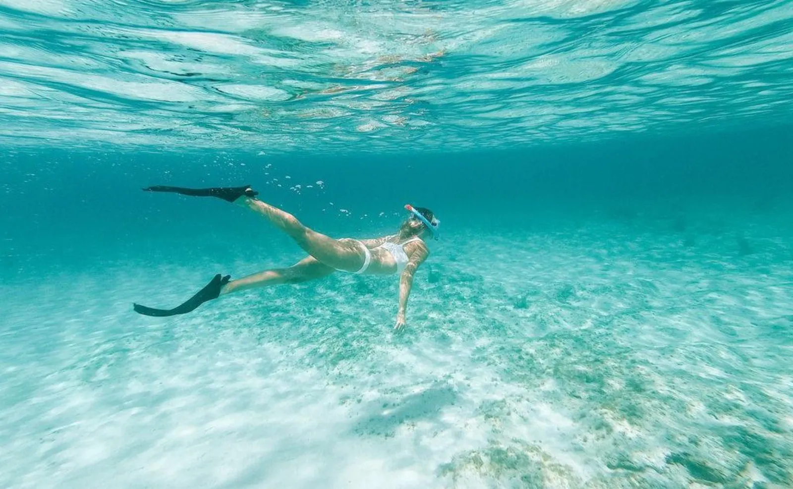 A swimmer in mask and fins under the surface, above a pale sandy bottom in turquoise water.