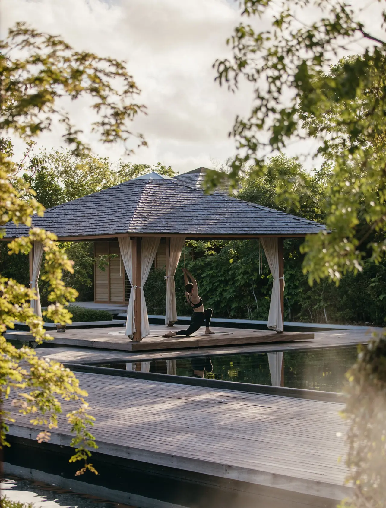 An open pavilion with pale curtains over a dark pond, where a guest practises yoga, among the spa’s trees.