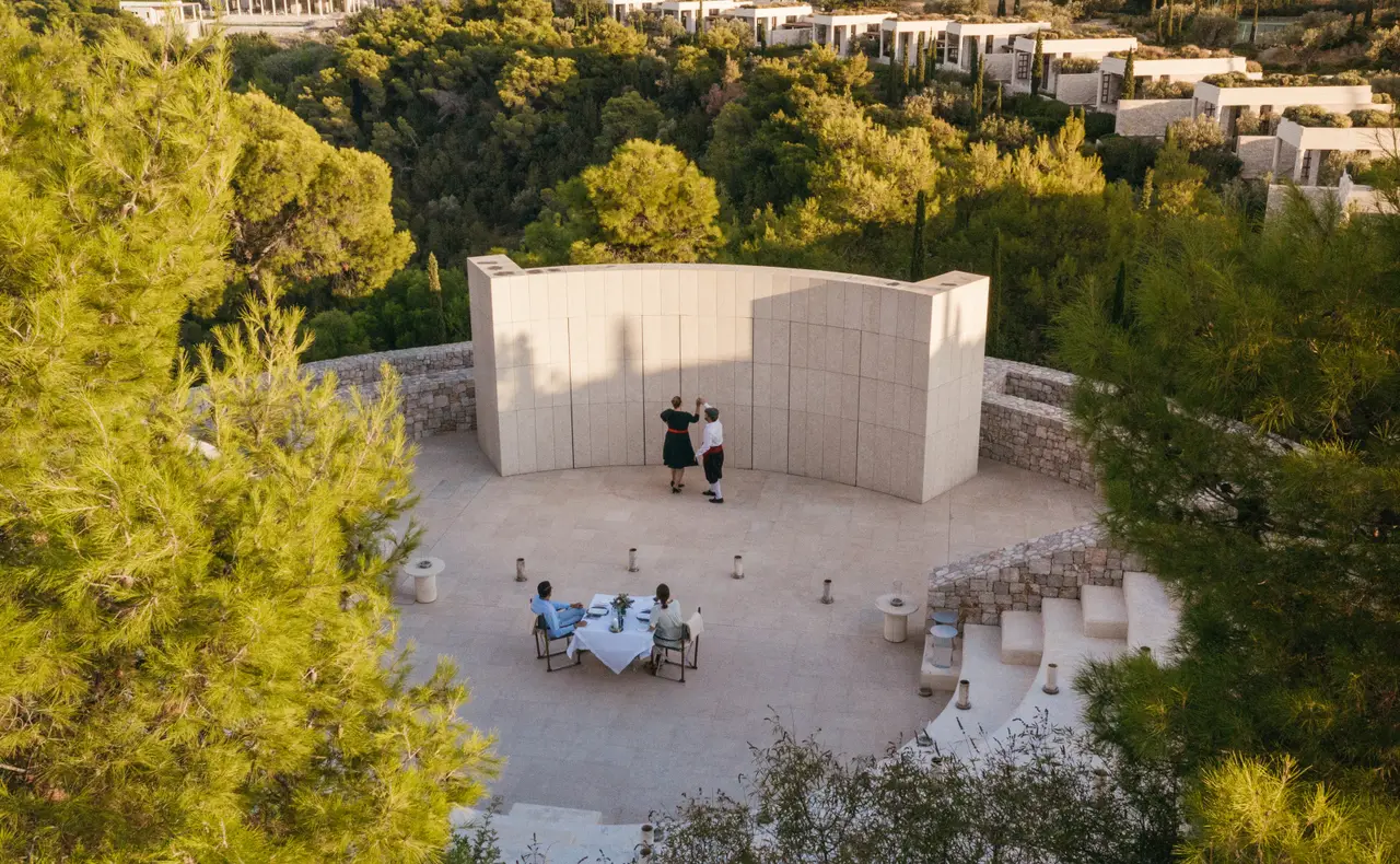 The open-air amphitheatre from above: 2 guests at a table in the middle of the stage and 2 figures standing before the curved stone wall, among pines.