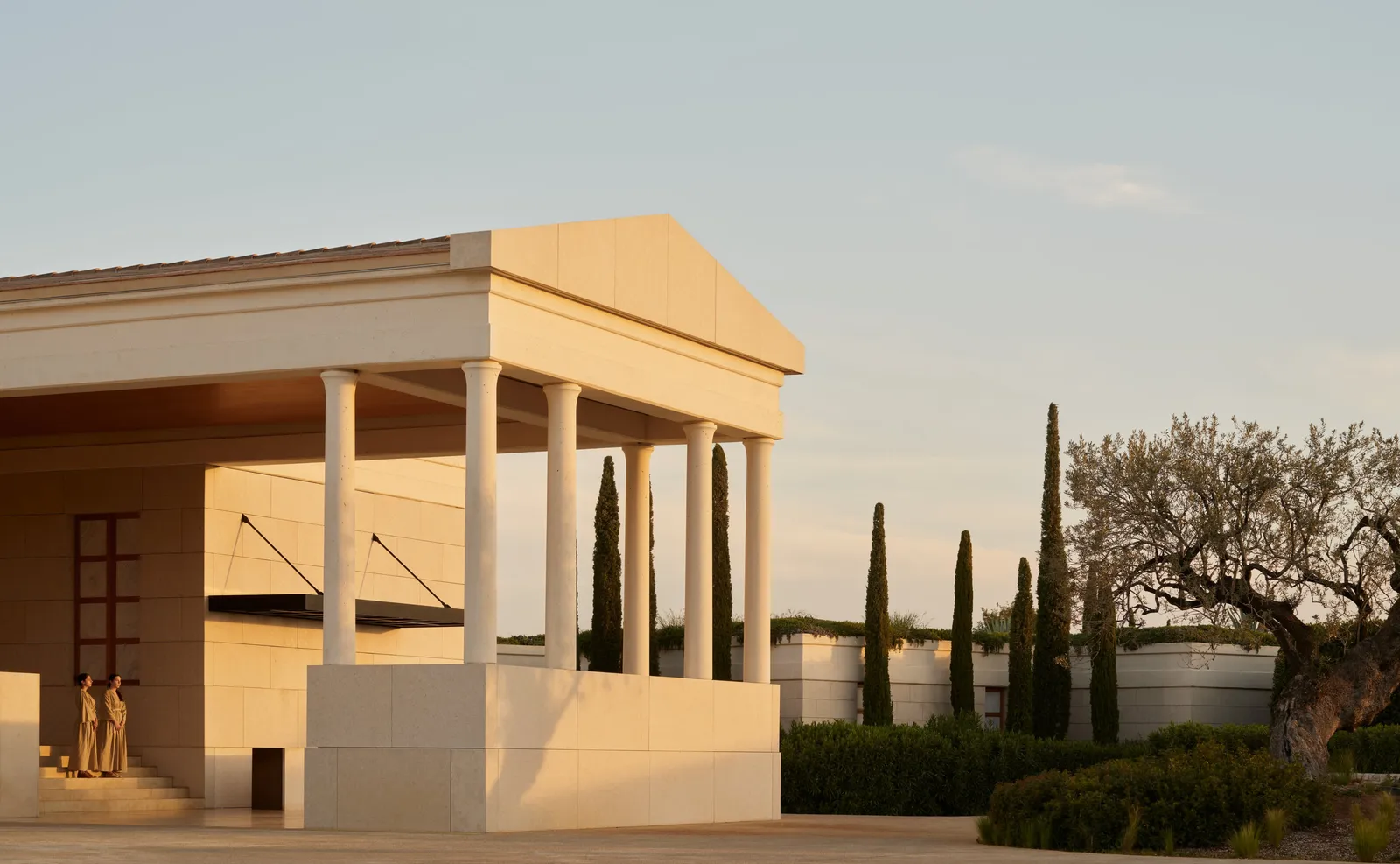 The arrival pavilion in golden light: pediment, columns, an old olive tree and a row of cypresses.