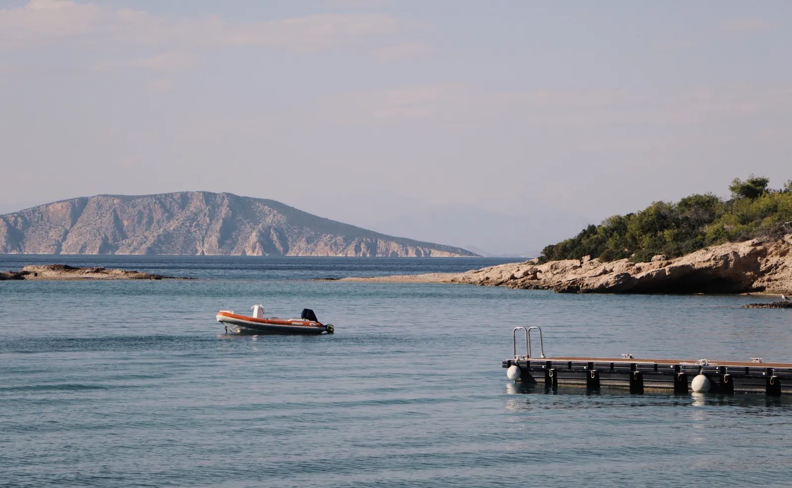 The Beach Club’s jetty with a dinghy moored off it, on calm water before a rocky coast.