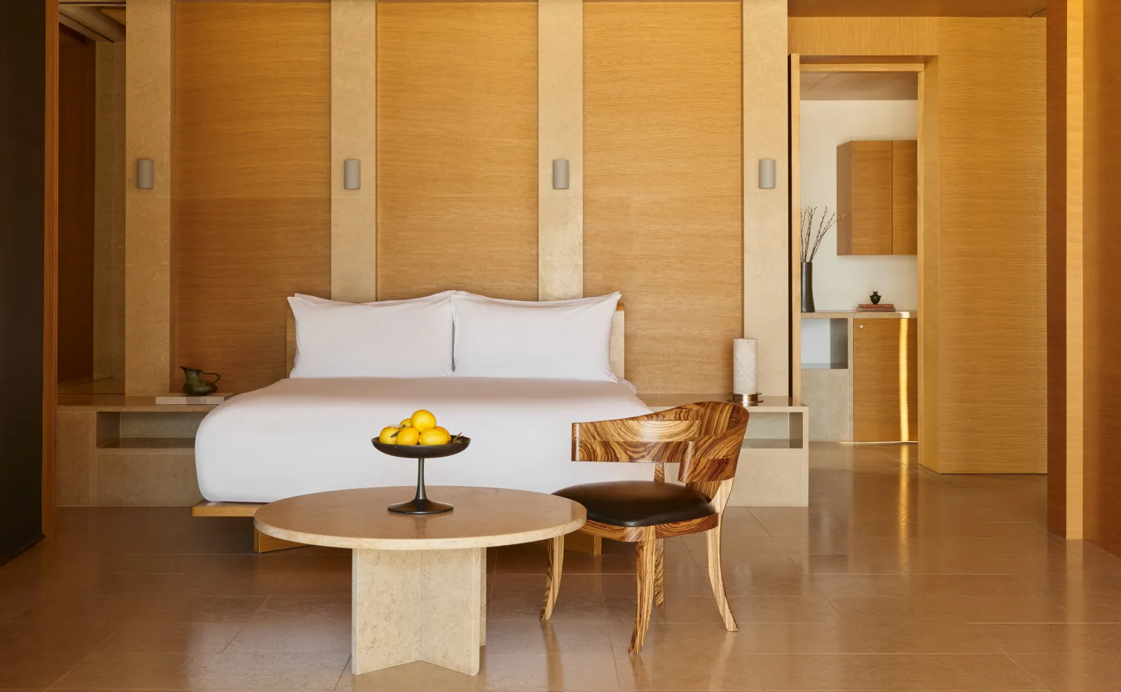 A Beach Pavilion’s bedroom: the white bed against a timber wall, a round stone table and a chair.