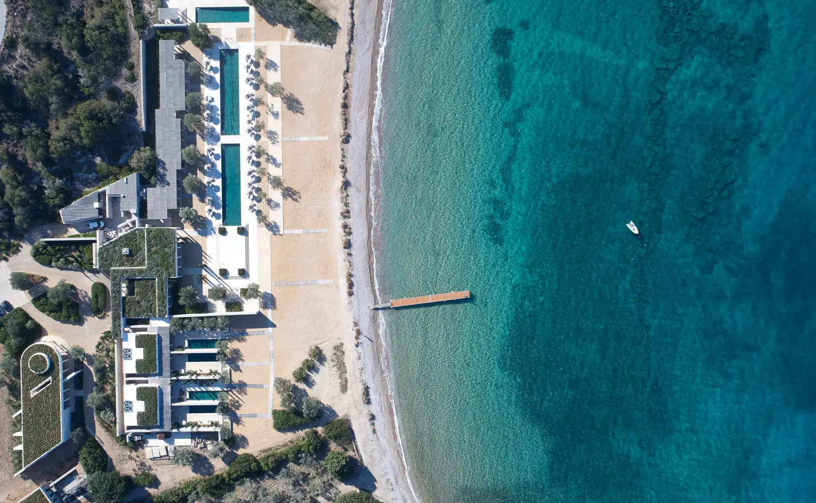 The Beach Club from above: long pools parallel to the shore, the pale beach, a jetty and turquoise sea.