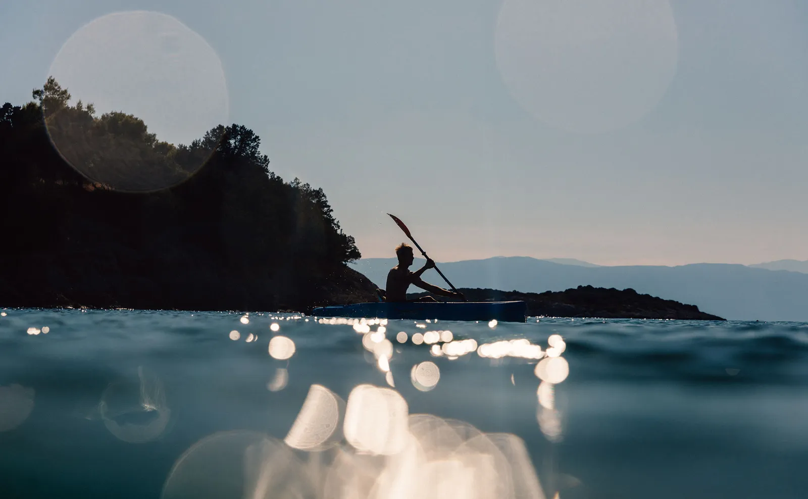 A kayaker in silhouette at dusk, the sun glittering on the water and a dark headland behind.