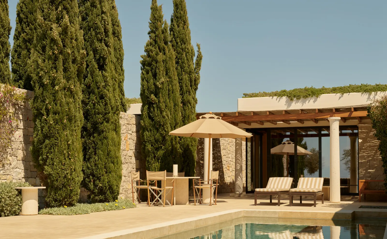 A Beach Pavilion: the pool before the pergola, a parasol, 2 loungers and cypresses against the stone wall.