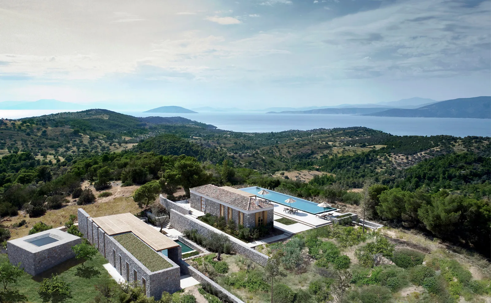 The one-bedroom villa from above: stone buildings with a green roof, a long pool and wooded hills running down to the sea.