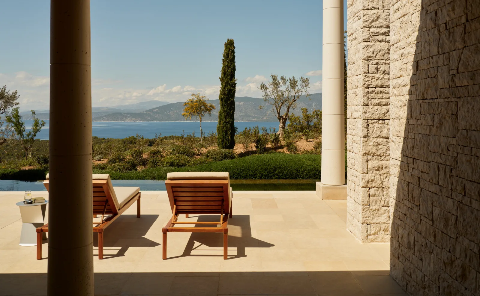 A sea-view pavilion’s terrace: 2 wooden loungers in the sun, a cypress and the gulf beyond the olive trees.