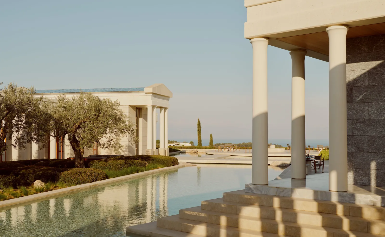 A colonnade with steps descending into a pool of water; beyond it a pedimented pavilion, olive trees and the sea.