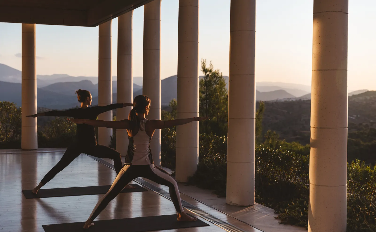 2 people in a yoga pose in the colonnaded pavilion, with the hills and the sea in early-morning light.