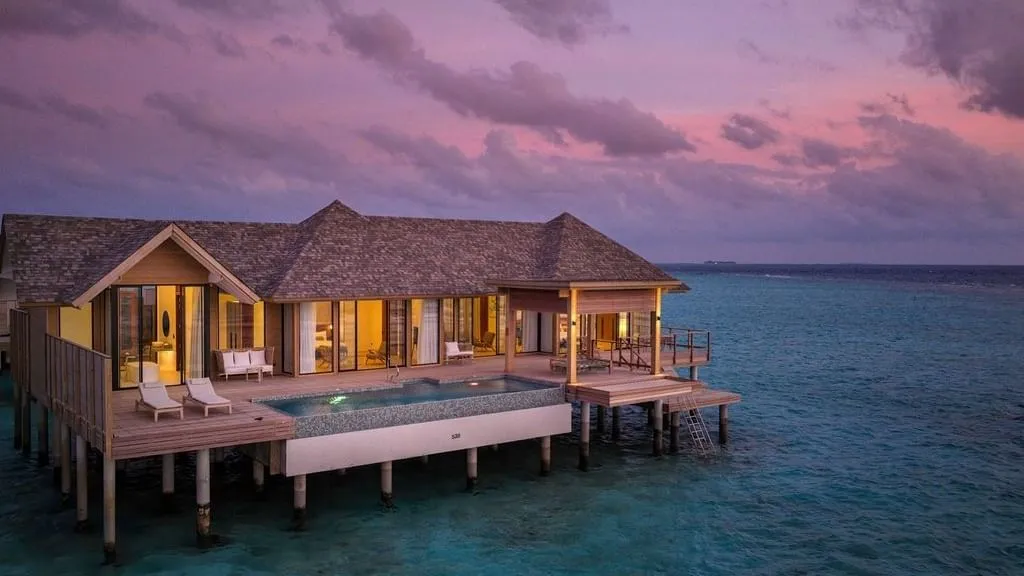 The Vilu residence over the water at dusk, its infinity pool and windows lit.