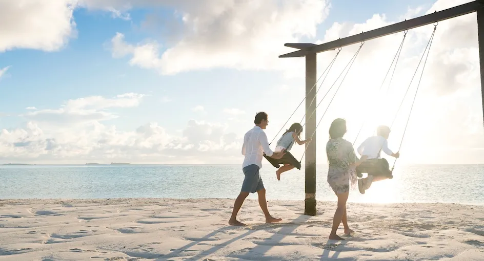 Una famiglia sull’altalena in spiaggia, con la laguna bassa e chiara alle spalle.