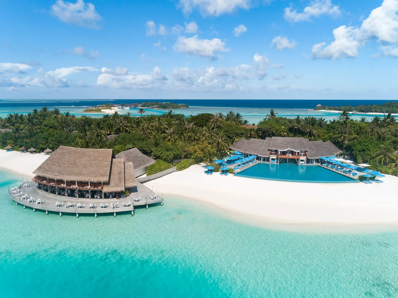 La piscina a sfioro sulla spiaggia, i lettini e gli ombrelloni blu, e a sinistra un padiglione di paglia sull’acqua.