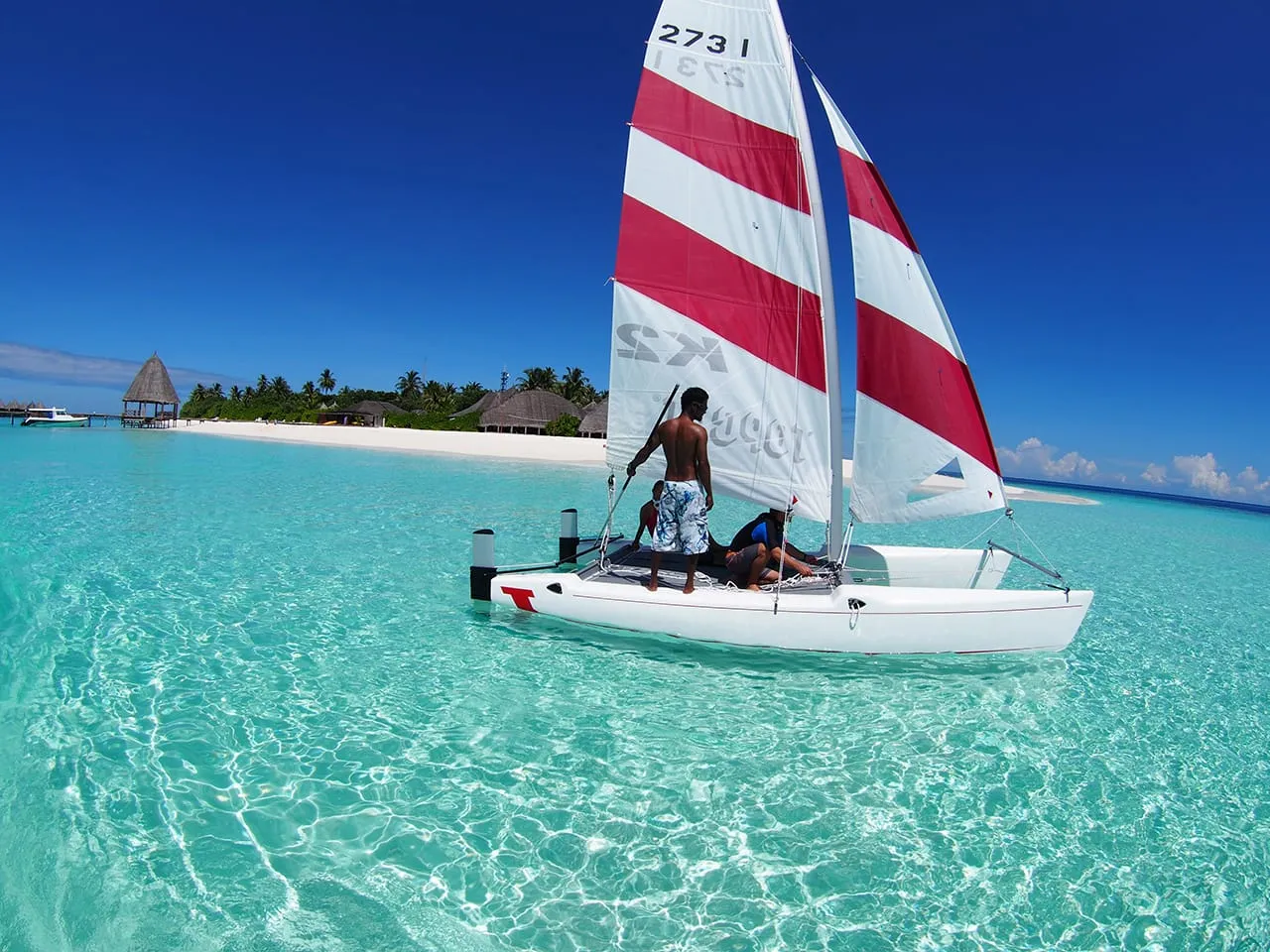 Un catamarano a vela bianco e rosso nella laguna trasparente, con l’isola sullo sfondo.