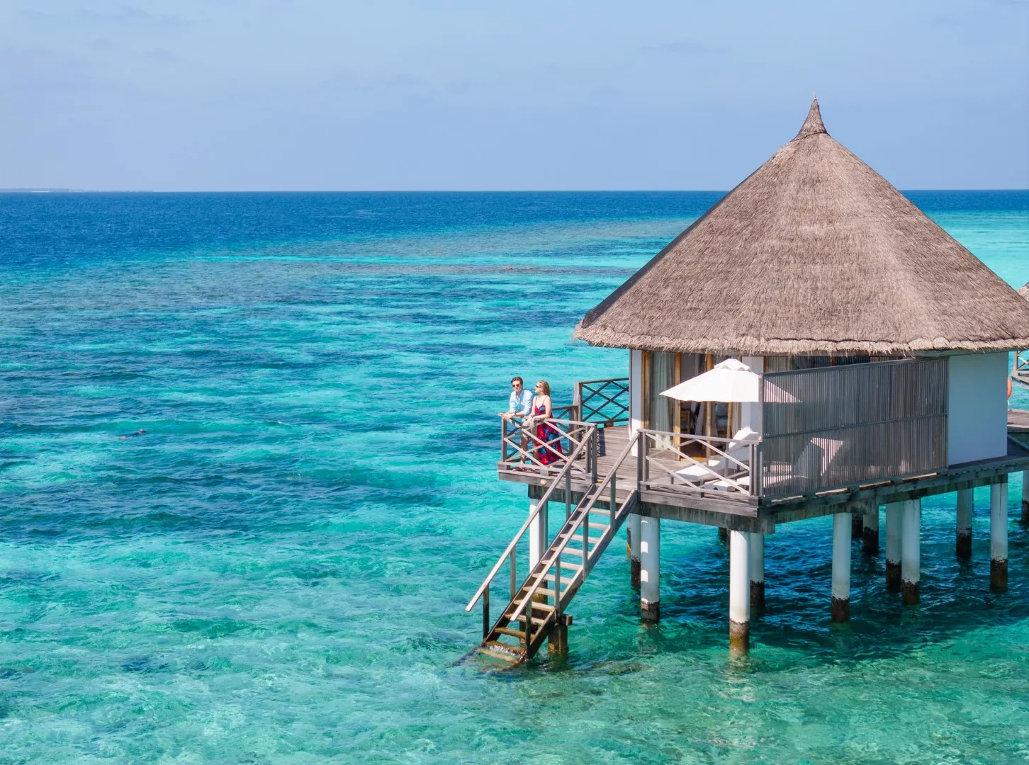 Un Superior Water Bungalow con il tetto di paglia sopra il reef, e una coppia sulla scaletta che scende in acqua.