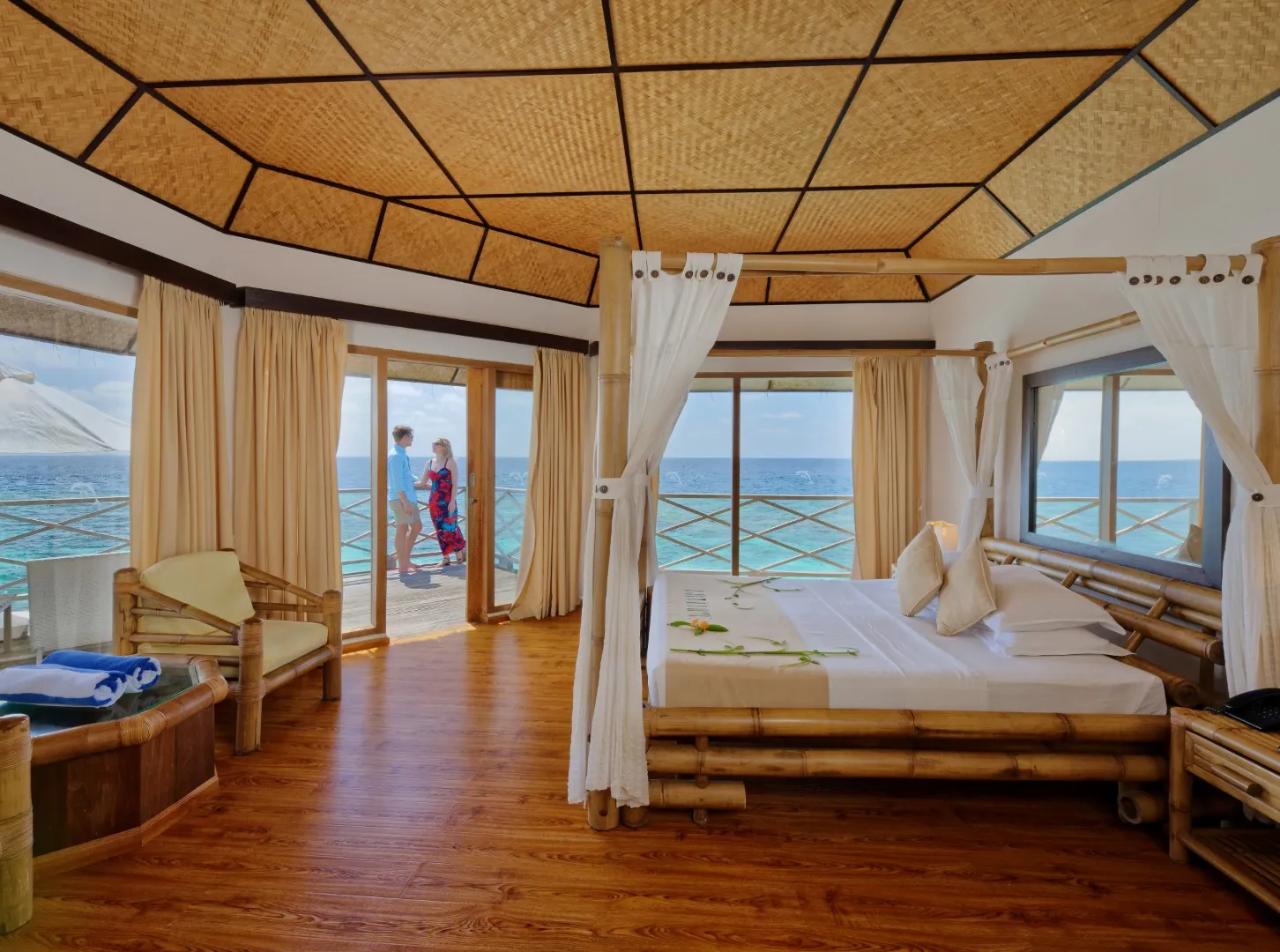 La camera di un Superior Water Bungalow, con il letto di bambù, il soffitto intrecciato e una coppia sulla terrazza.