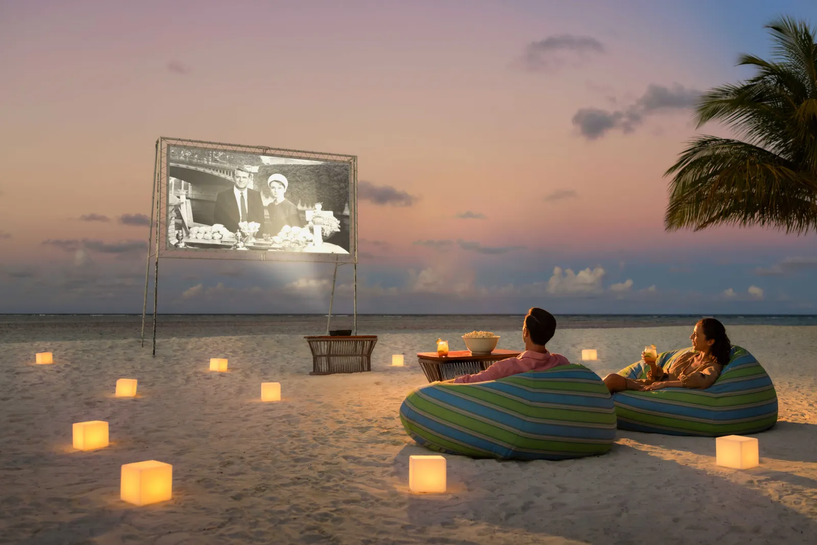 A cinema on the beach at sunset: the screen, 2 bean bags and lanterns lit on the sand.