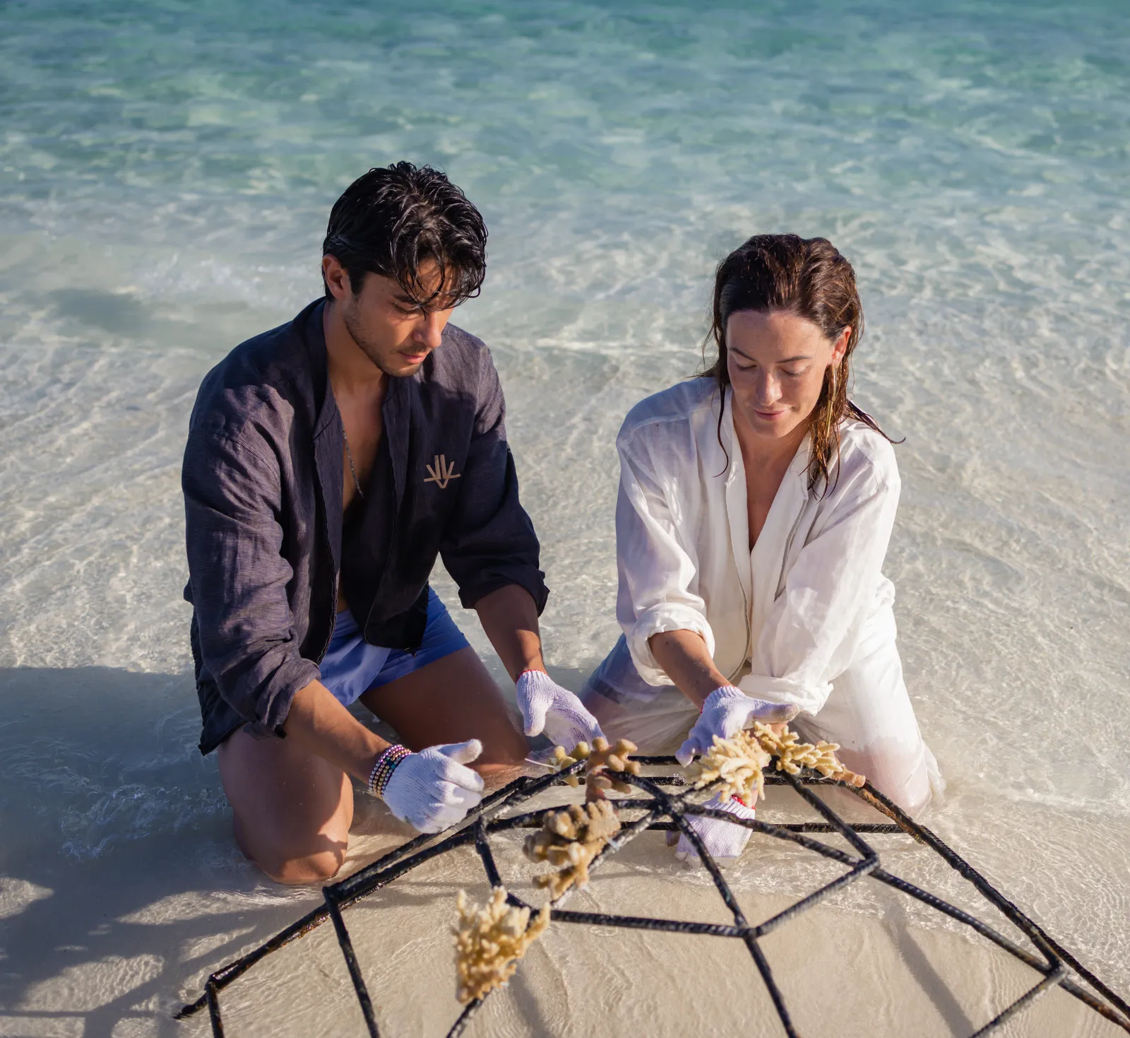 A couple tie coral fragments to a metal frame, kneeling in the shallows.