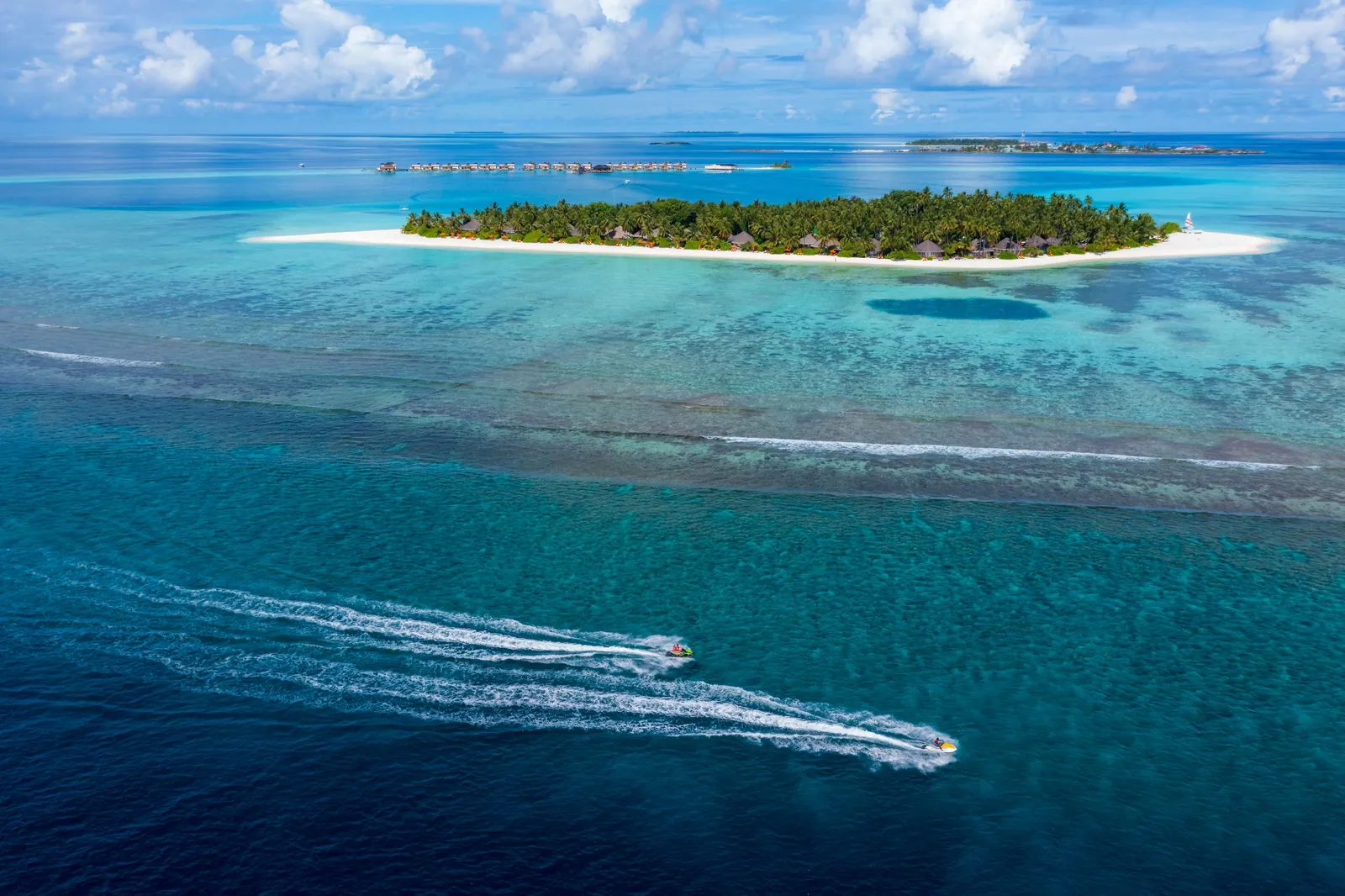 Velavaru from above: the green island and its white beach in the lagoon, the water villas on the horizon and the wakes of 2 boats.
