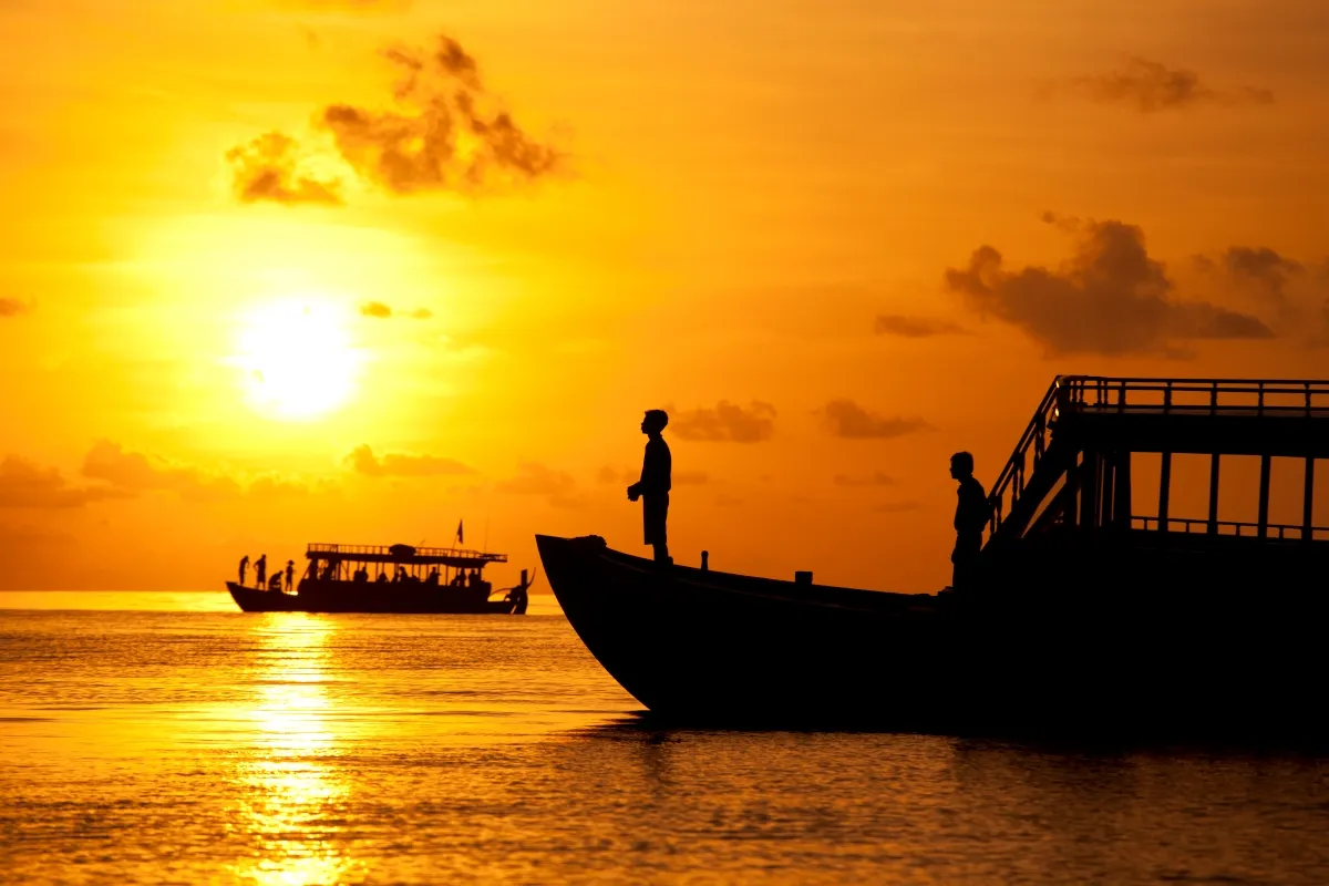 A dhoni in silhouette at sunset, a figure standing at the bow and the sun on the water.