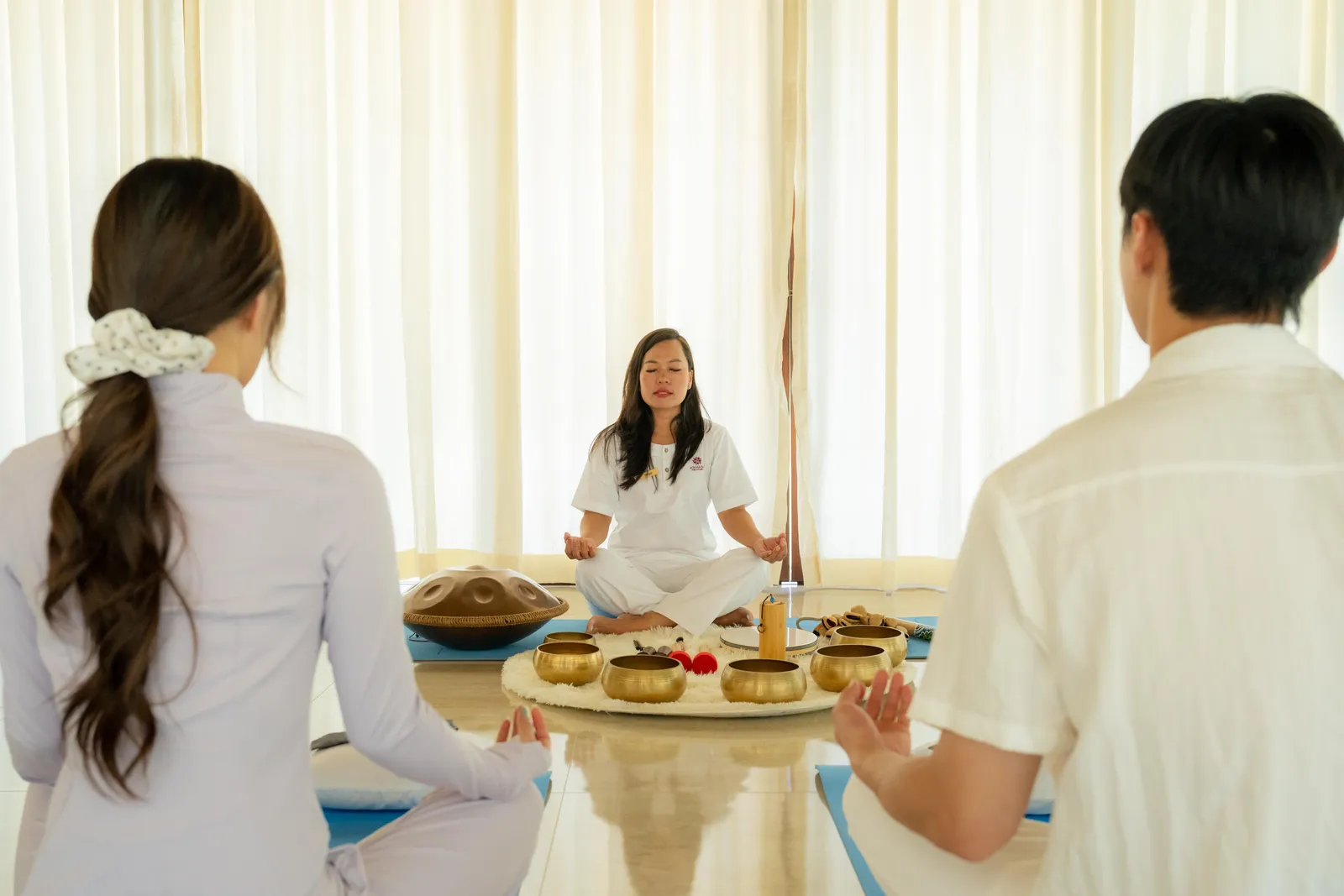 A singing-bowl session in a bright room, the teacher seated before 2 guests.
