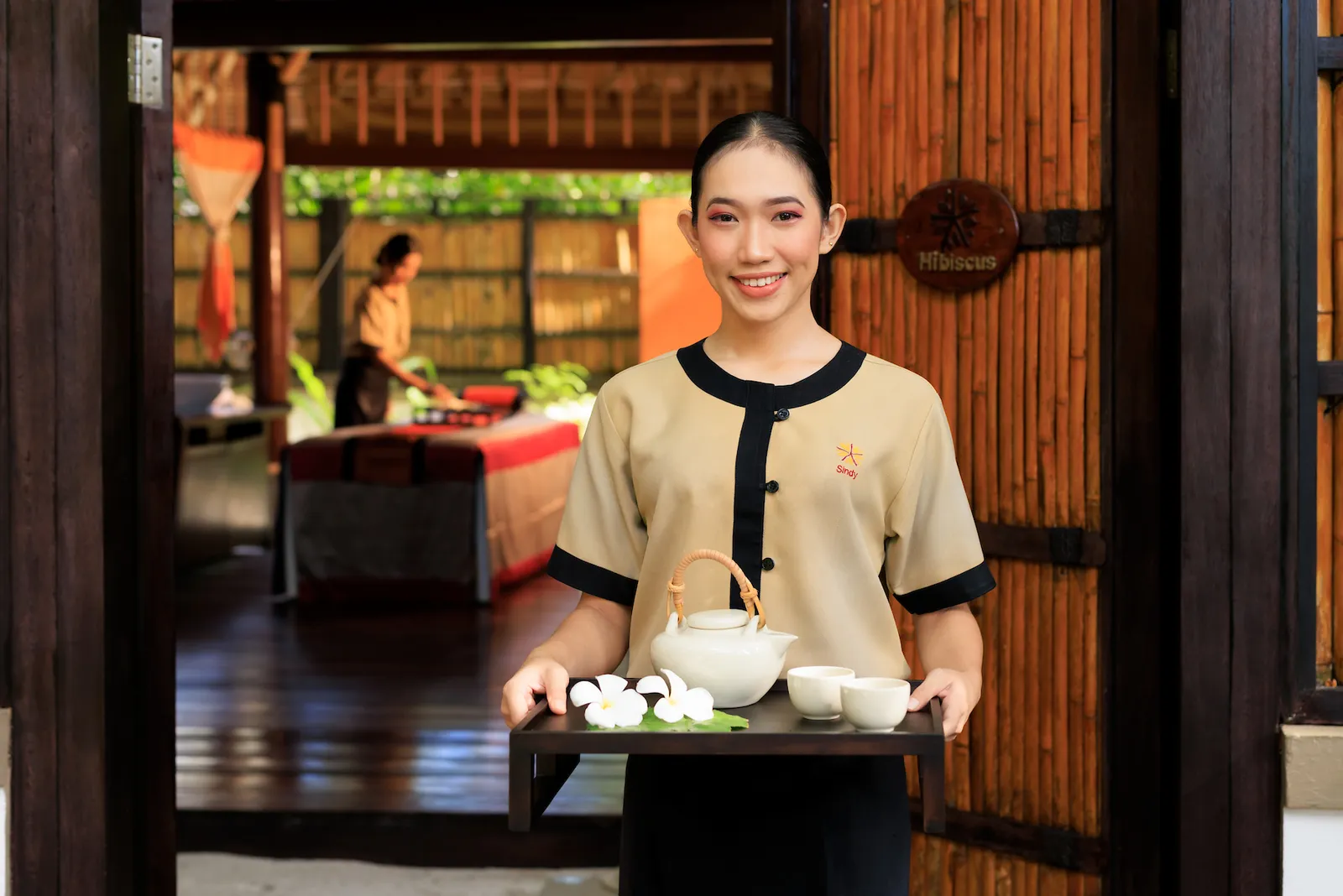 A therapist smiles at the spa’s door with a tray of tea, a treatment pavilion behind her.