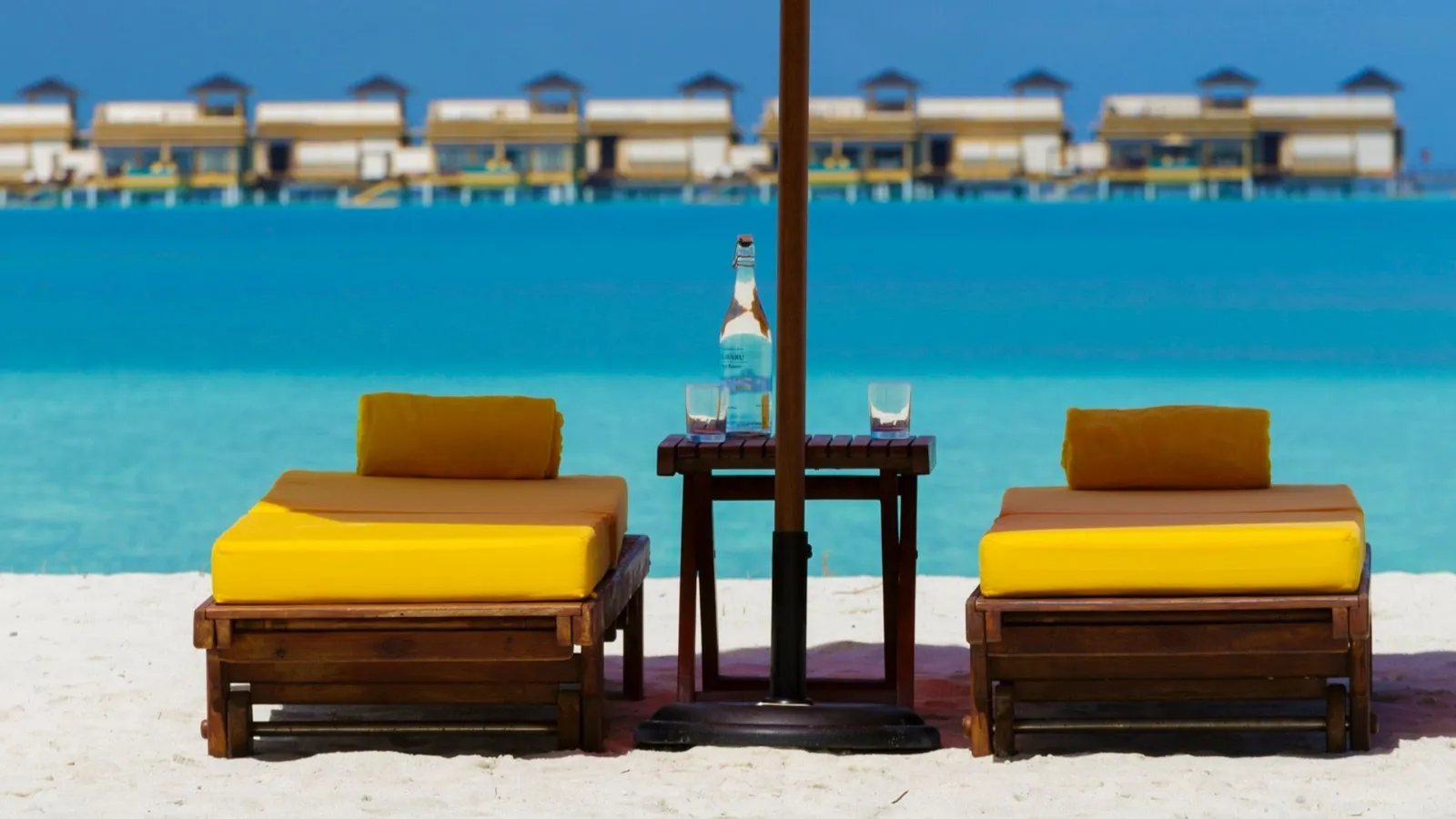 2 yellow loungers under a parasol on the beach, with the line of water villas across the lagoon.