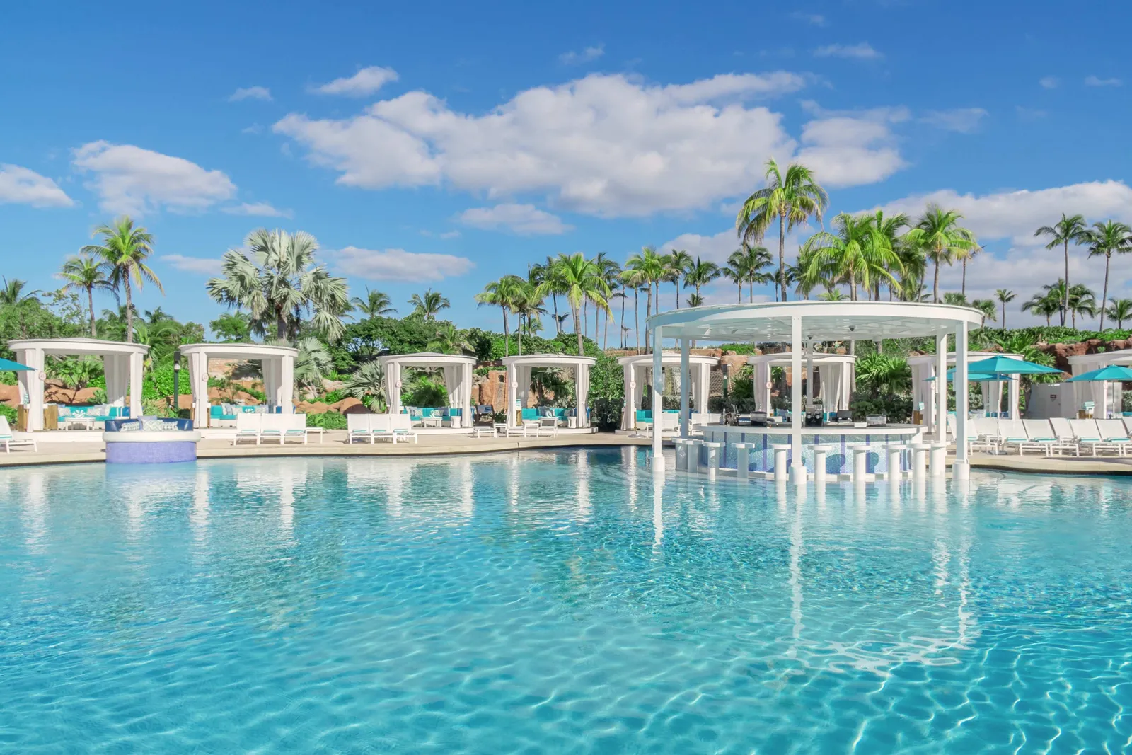 A wide blue pool ringed by white curtained cabanas, with a swim-up bar and palms behind.
