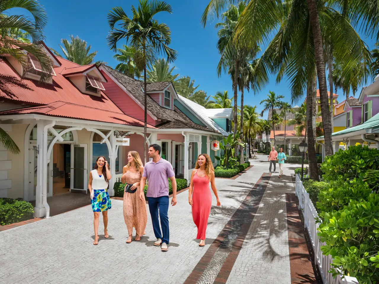 A pedestrian street between pastel-painted, red-roofed cottages and palms, with 4 people strolling.