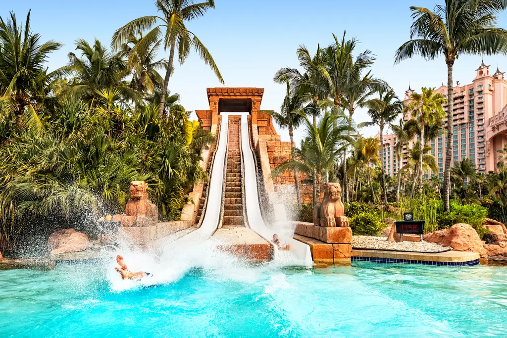 2 slides run down the Mayan Temple pyramid between palms; at the bottom a swimmer lands in the pool in a burst of spray.