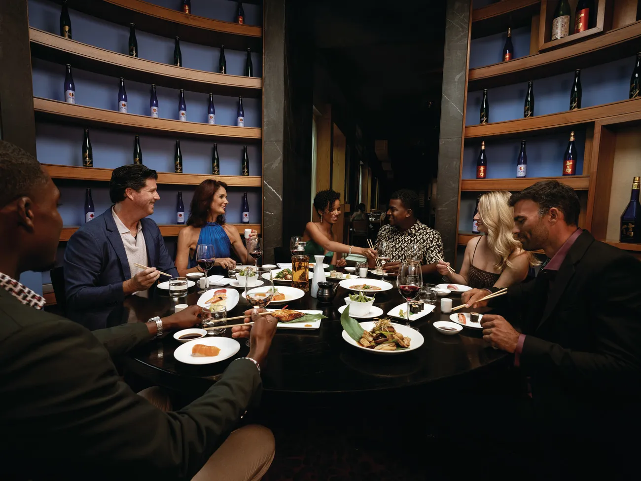 A table of 8 at dinner, plates of sushi in the middle and walls of sake bottles lit in blue behind them.